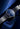 A close-up of a wristwatch with a dark blue dial, silver-toned case, and matching blue strap, photographed against a dark blue abstract background.